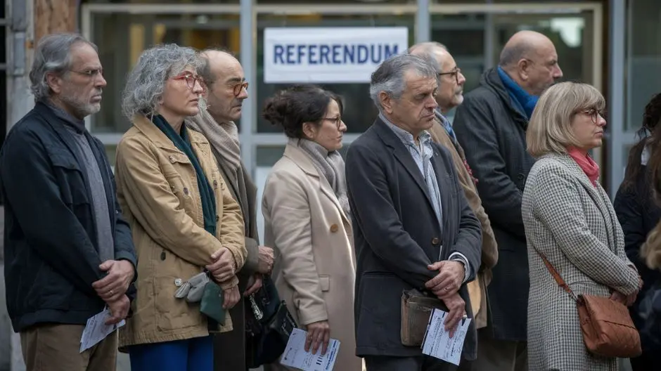Persone in fila davanti a un seggio elettorale per votare al referendum. Uomini e donne di ogni età, vestiti in modo semplice, con espressioni serie e attente. Alcuni tengono la tessera elettorale in mano.